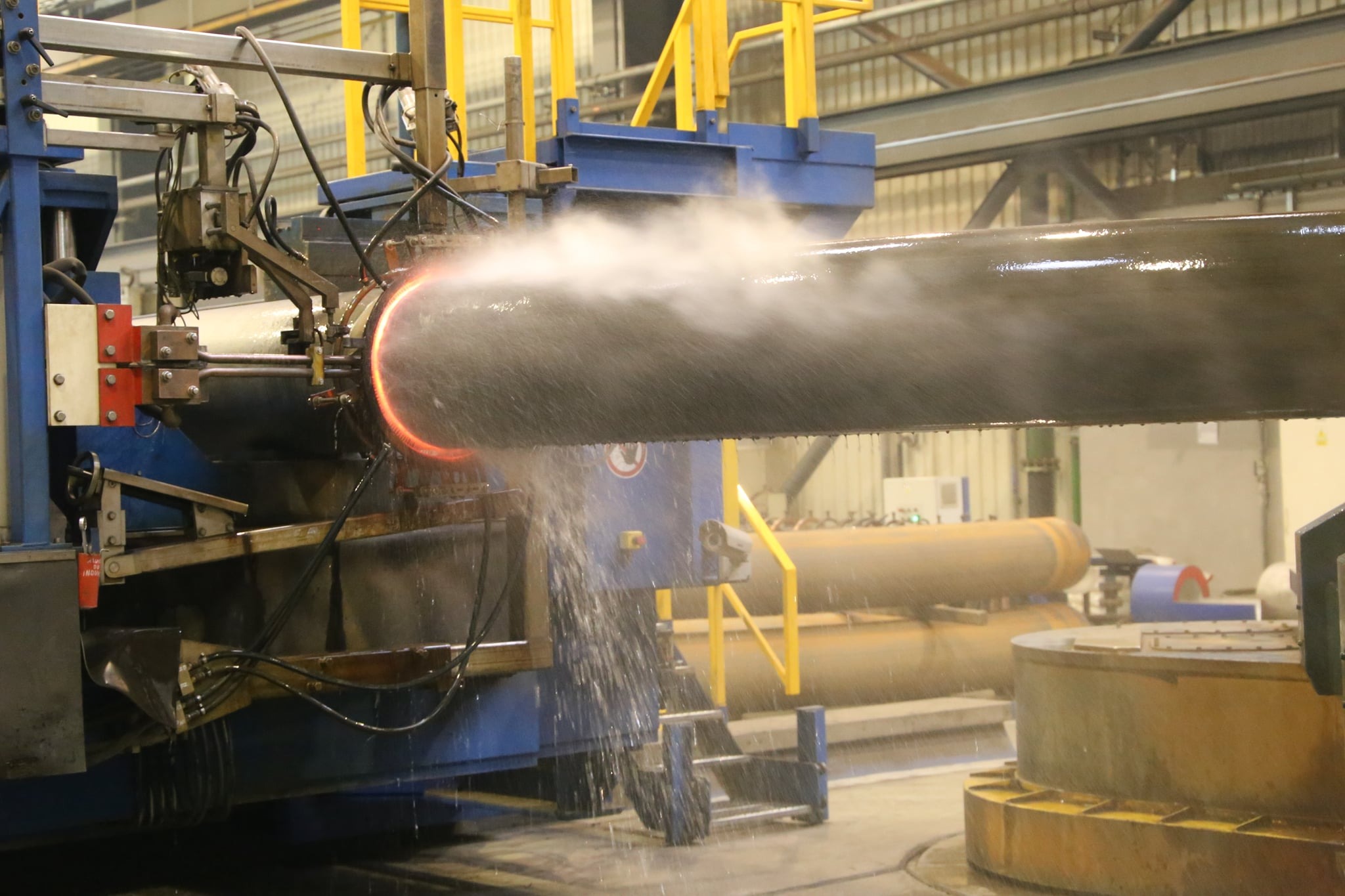 Proces obróbki termicznej rury przemysłowej za pomocą specjalistycznej maszyny, widać efekt nagrzewania i chłodzenia wodą, generujący parę.