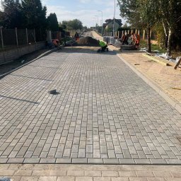 Wnortex Construction Sp. z o.o. - Ułożona kostka brukowa na długiej drodze z pracownikami w tle. Szara kostka, jasne niebo, drzewa po bokach. Etap prac brukarskich w słoneczny dzień.