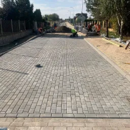 Ułożona kostka brukowa na długiej drodze z pracownikami w tle. Szara kostka, jasne niebo, drzewa po bokach. Etap prac brukarskich w słoneczny dzień.