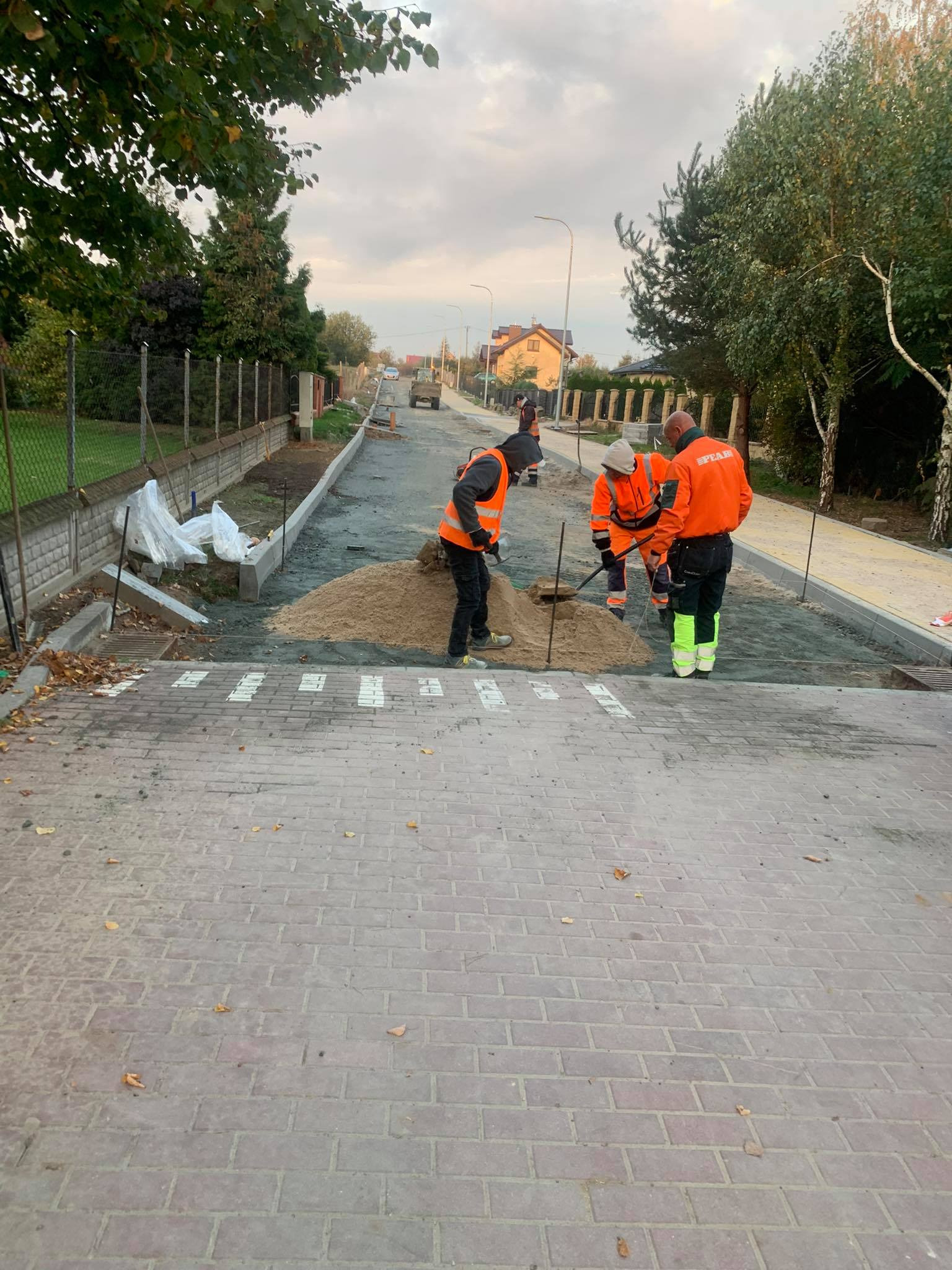 Robotnicy w odblaskowych ubraniach równają piasek pod nową nawierzchnię chodnika z kostki brukowej. W tle widoczny krajobraz miejski z drzewami i budynkami.
