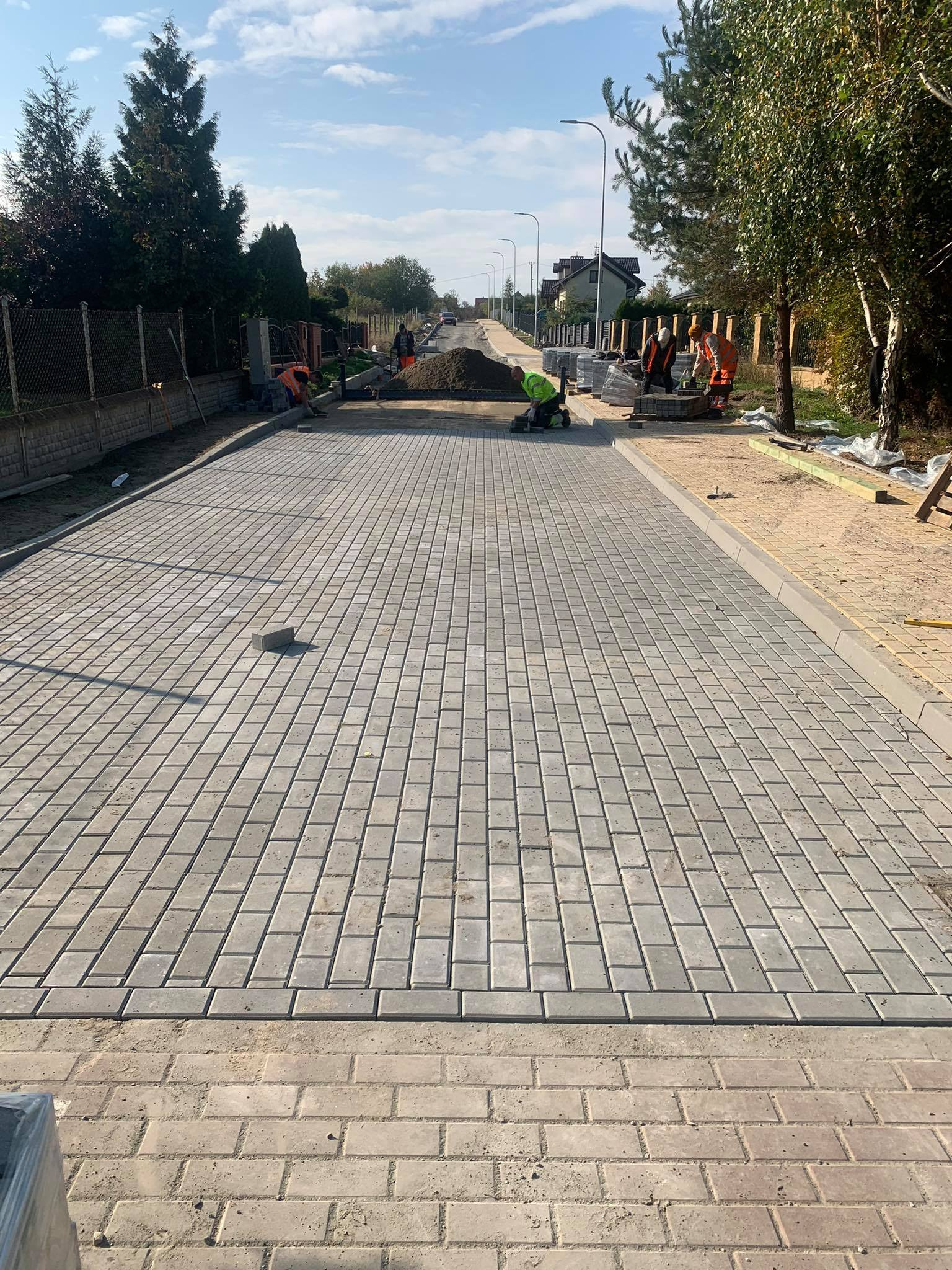 Ułożona kostka brukowa na długiej drodze z pracownikami w tle. Szara kostka, jasne niebo, drzewa po bokach. Etap prac brukarskich w słoneczny dzień.