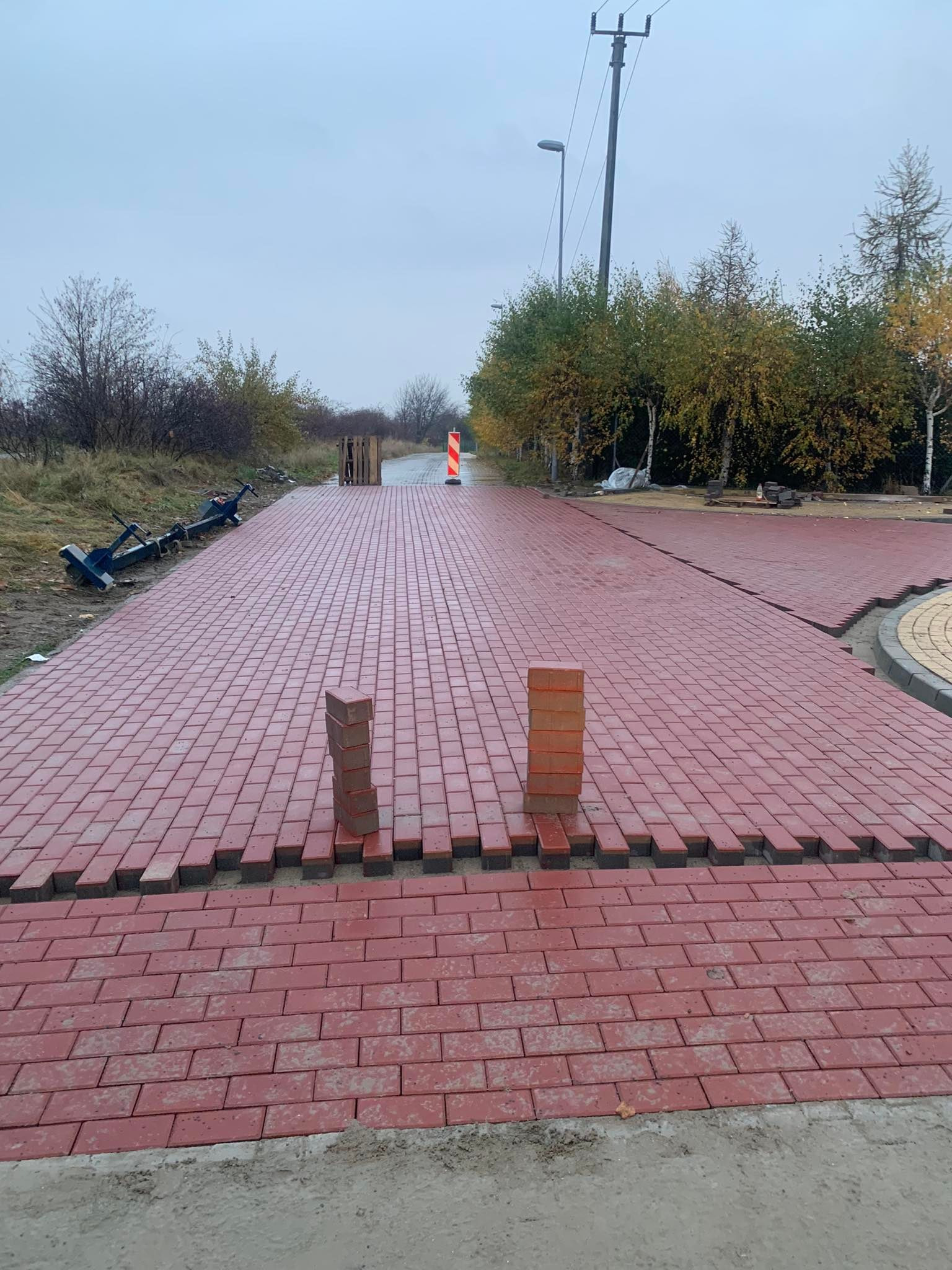 Czerwona kostka brukowa w trakcie układania na drodze, widoczne narzędzia i paleta w tle. Częściowo ułożona nawierzchnia w jesiennej scenerii.