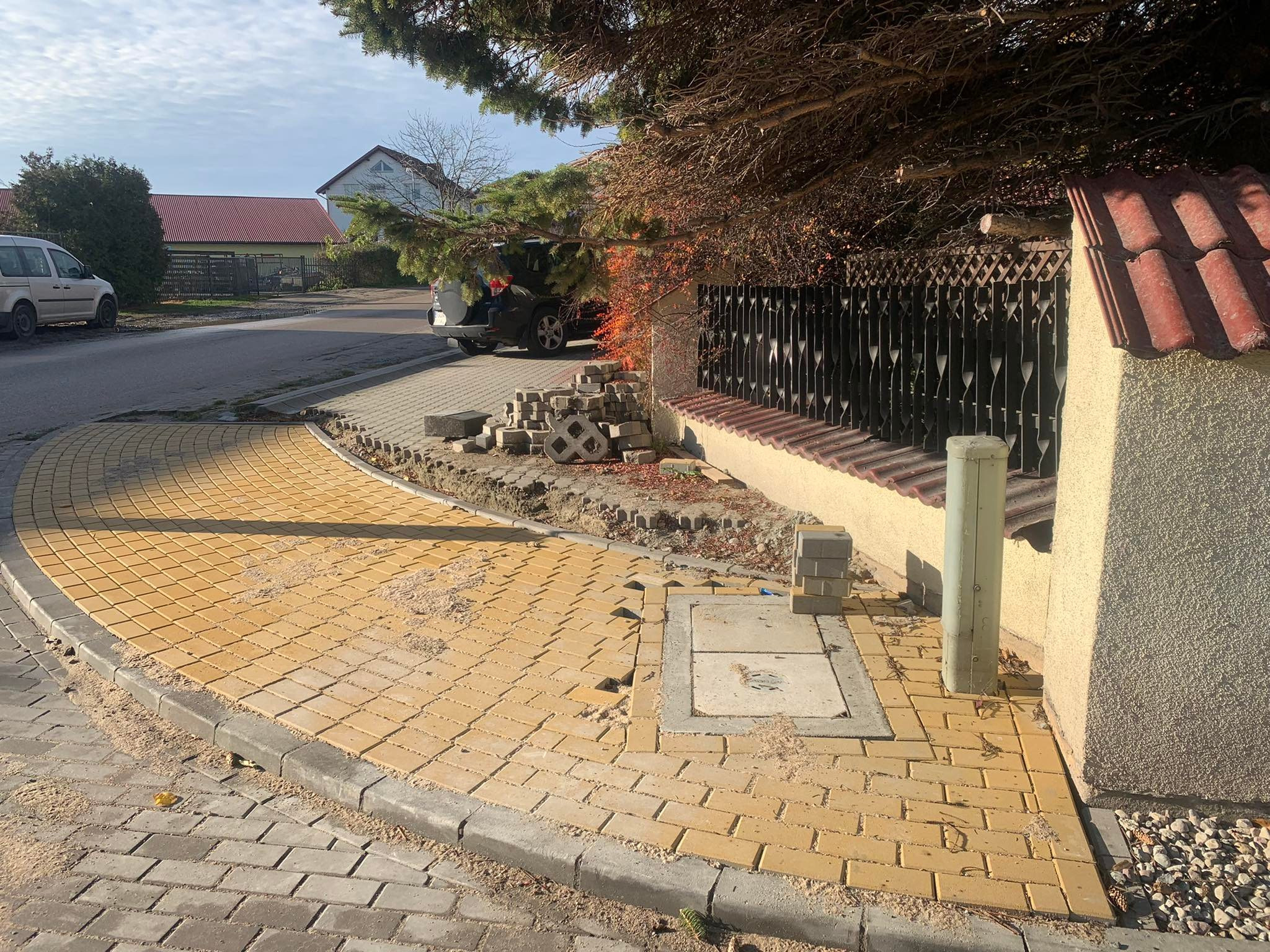 Żółta kostka brukowa ułożona na zakręcie chodnika, z widocznym włazem studzienki i przygotowanym miejscem na dalsze prace brukarskie przy ogrodzeniu.