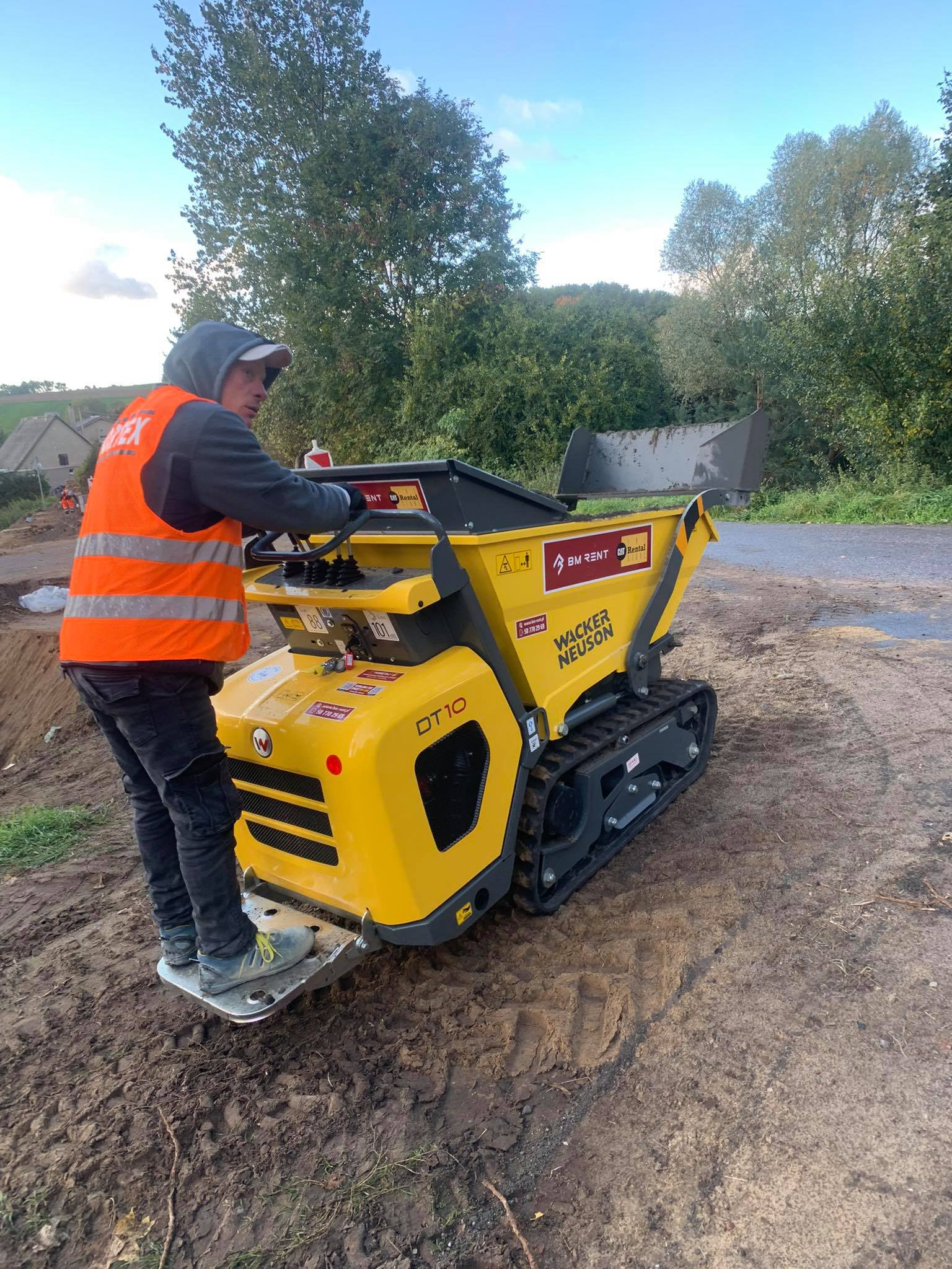 Pracownik w kamizelce obsługuje żółtą mini-ładowarkę gąsienicową Wacker Neuson DT10 na placu budowy. W tle drzewa i budynki.