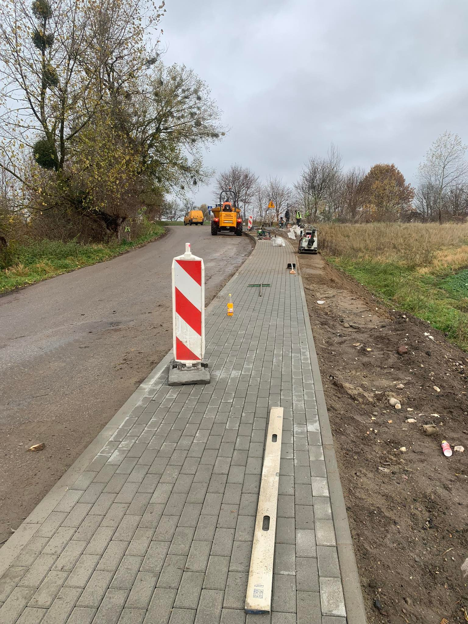 Układanie kostki brukowej na poboczu drogi, widoczny poziomica i maszyny budowlane w tle. Prace w toku, jesienna sceneria.