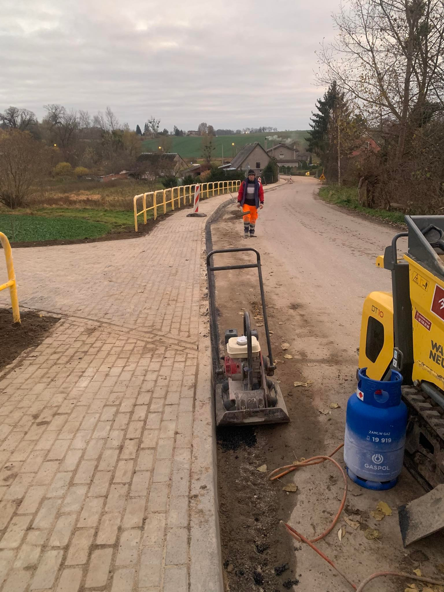 Układanie kostki brukowej na poboczu drogi w terenie wiejskim. Widoczny pracownik w odblaskowej odzieży i maszyny budowlane. Żółte barierki zabezpieczają teren prac.