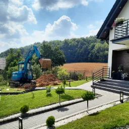 Niebieska koparka na tle nowoczesnego domu z balkonem i zadbanym ogrodem, w tle pole i las. Wykopana ziemia obok, widoczny zbiornik na wodę.