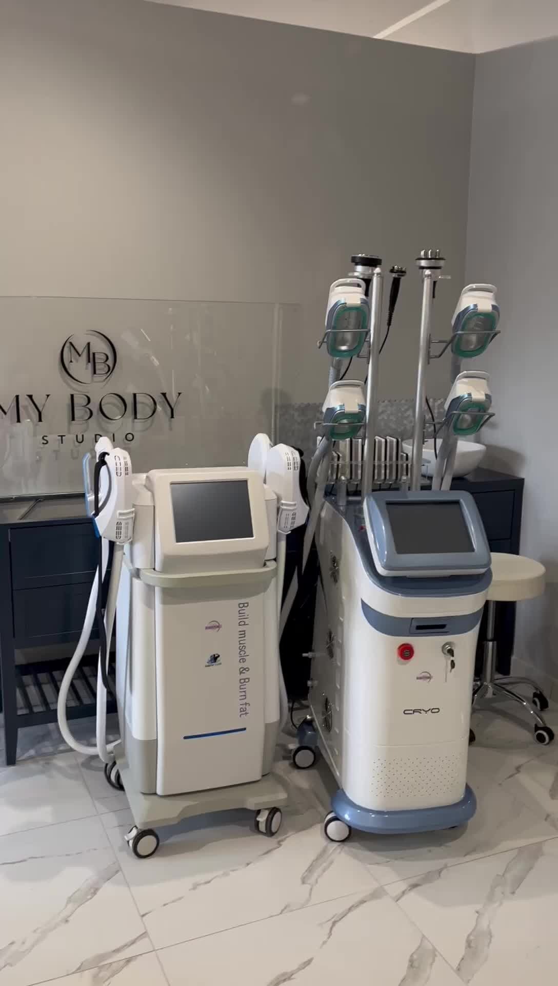 Dwa nowoczesne urządzenia do modelowania sylwetki w salonie kosmetycznym, jedno z napisem 'Build muscle & Burn fat', drugie 'CRYO', na tle szklanej ściany z logo 'My Body Studio'.