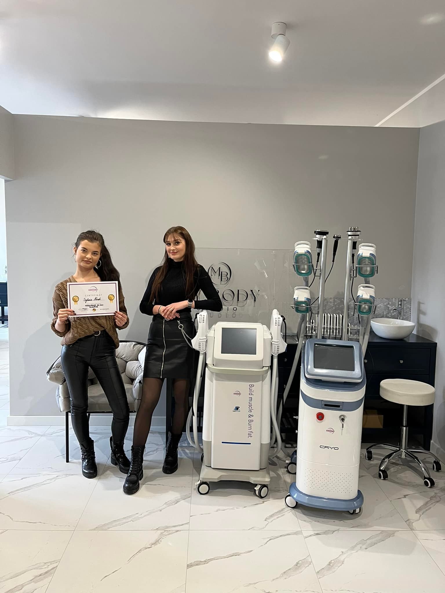 Dwie kobiety prezentują certyfikat ukończenia szkolenia z zakresu modelowania sylwetki, stojąc obok profesjonalnych urządzeń do kriolipolizy i elektrostymulacji w nowoczesnym gabinecie kosmetycznym...