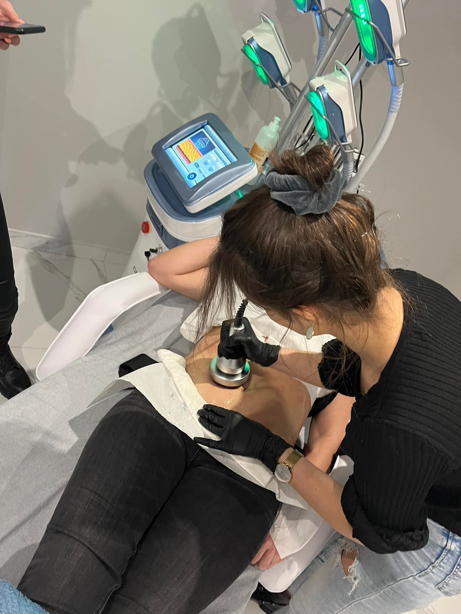 Kobieta w czarnych rękawiczkach wykonuje zabieg na brzuchu klientki leżącej na białym prześcieradle, używając urządzenia z podświetlanymi głowicami i ekranem dotykowym.