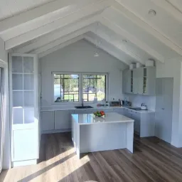 Jasna kuchnia z wyspą i drewnianą podłogą w domu z skośnym sufitem. Otwarta szafka z przeszkleniami po lewej. Za oknem widoczny teren zielony.