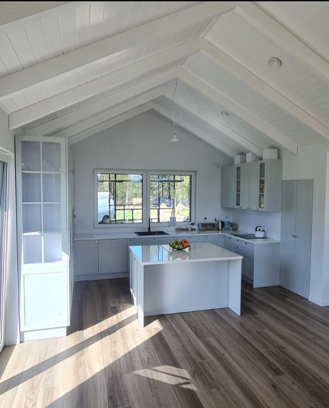 Jasna kuchnia z wyspą i drewnianą podłogą w domu z skośnym sufitem. Otwarta szafka z przeszkleniami po lewej. Za oknem widoczny teren zielony.