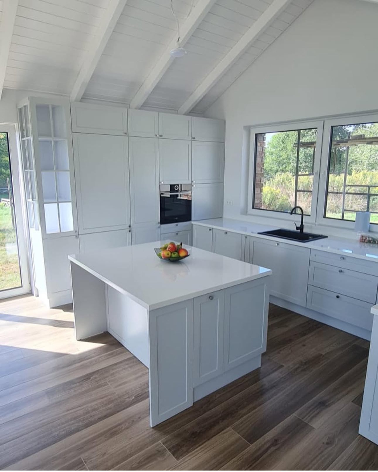 Jasna kuchnia z wyspą i drewnopodobną podłogą. Duże okno z widokiem na zieleń. Na wyspie misa z owocami. Minimalistyczny styl z białymi szafkami.