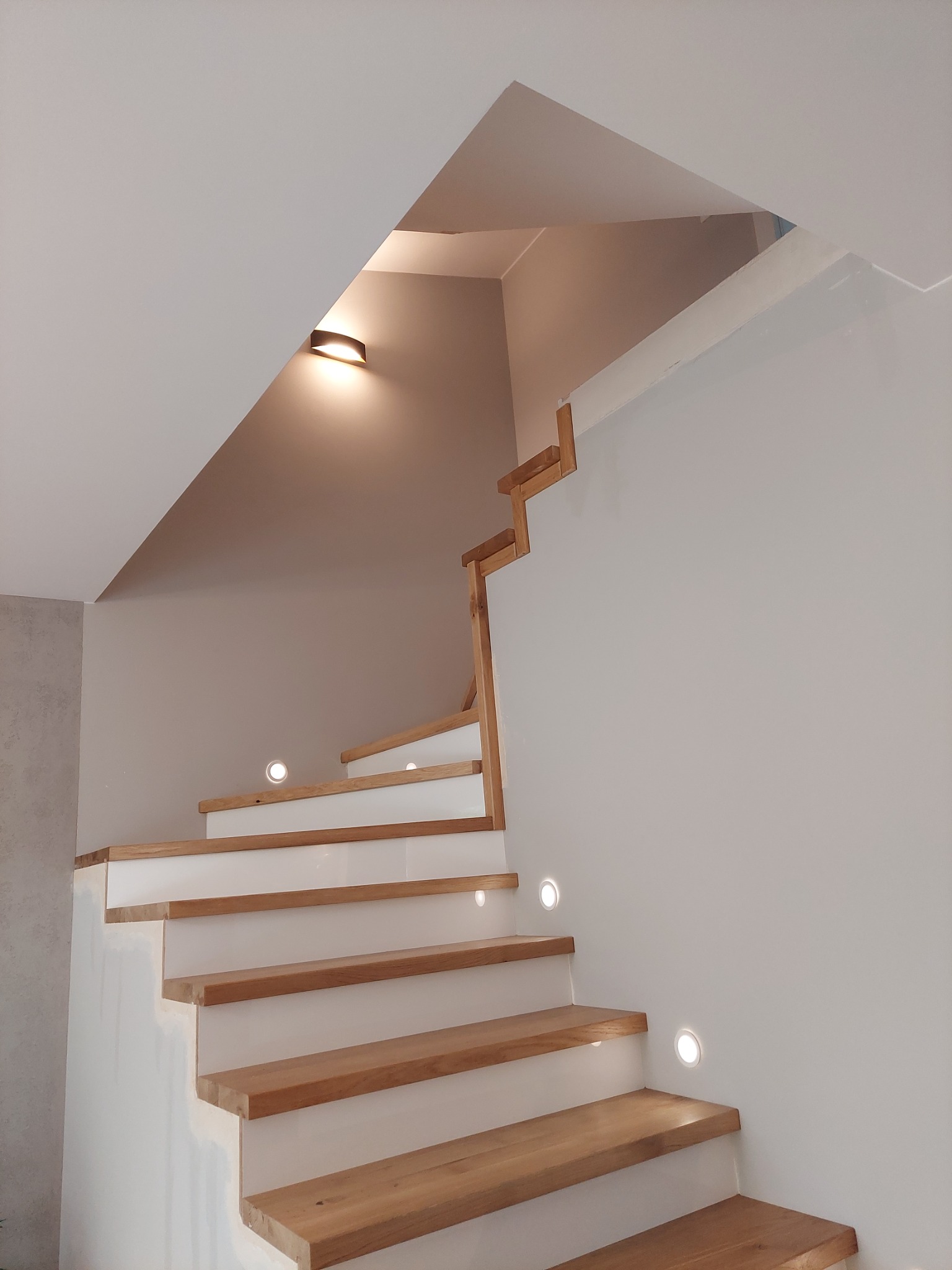 Nowoczesne schody drewniane z białymi podstopnicami, oświetlone punktowymi lampami LED w ścianie i lampą sufitową, minimalistyczny design.
