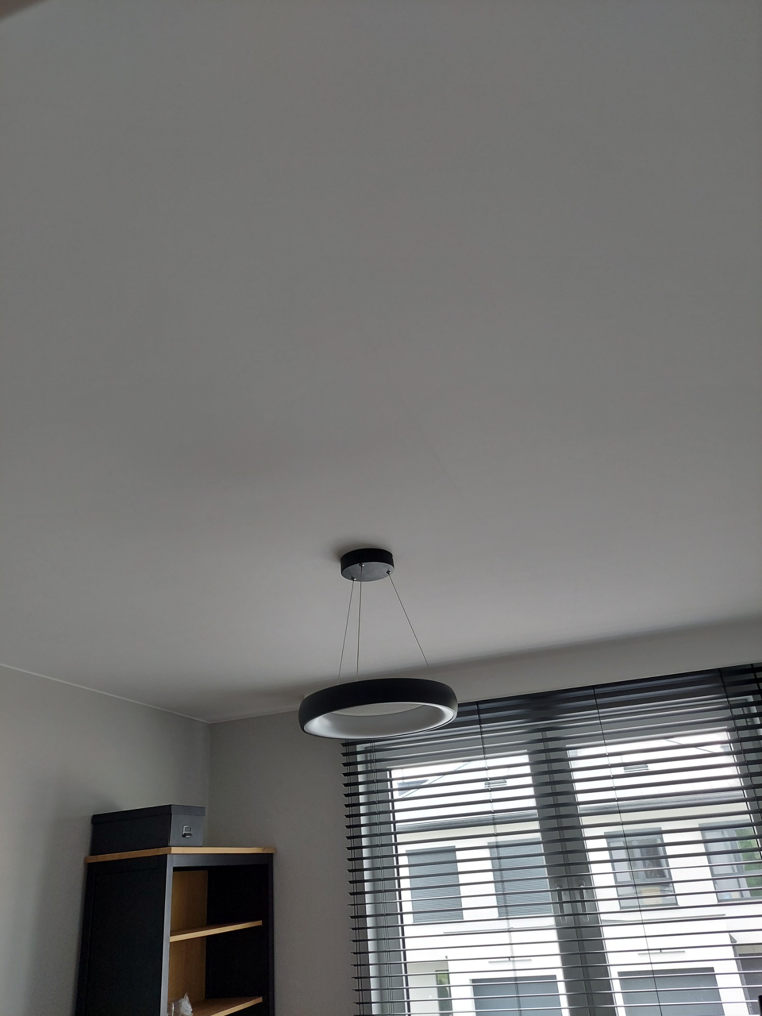 Czarno-biała okrągła lampa wisząca z widoczną konstrukcją mocującą, na tle okna z żaluzjami i regału z pudełkiem na górze.