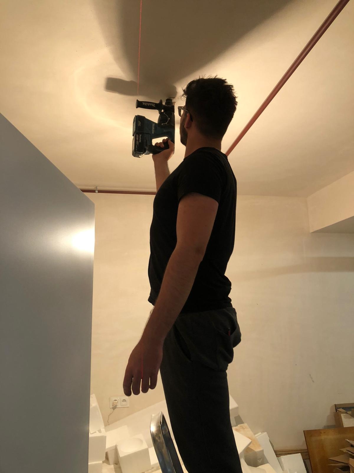 Mężczyzna w okularach, stojąc na drabinie, wierci wiertarką akumulatorową Makita otwór w suficie, pod czerwoną rurą biegnącą wzdłuż sufitu. Na podłodze widoczne białe płyty styropianowe.