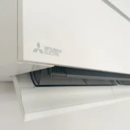 Biała jednostka wewnętrzna klimatyzatora Mitsubishi Electric zamontowana na ścianie, widoczna z bliska, z otwartą klapką nawiewu.