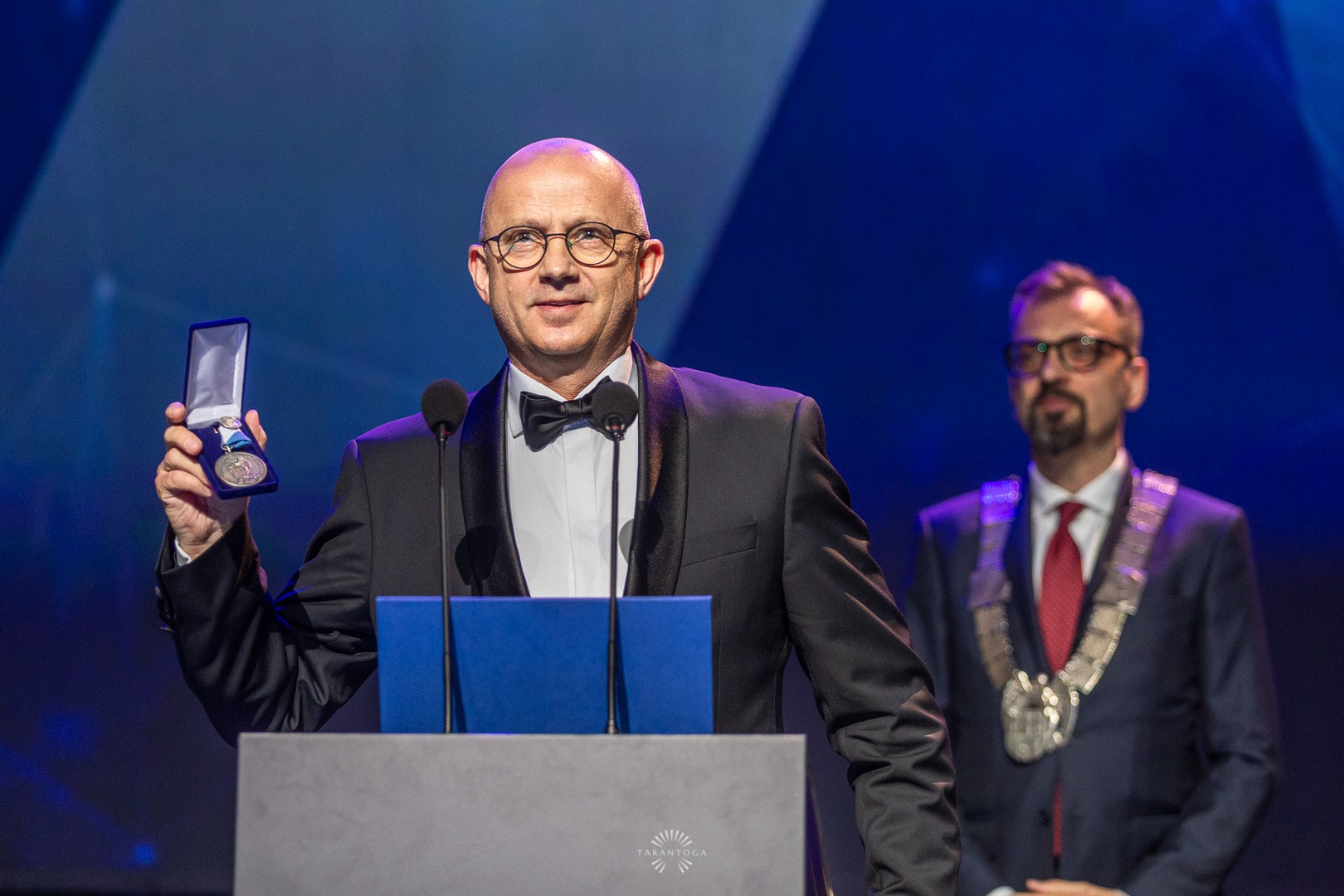 Mężczyzna w smokingu na scenie, trzyma w ręku otwarte pudełko z medalem, drugi mężczyzna w tle z łańcuchem na szyi.