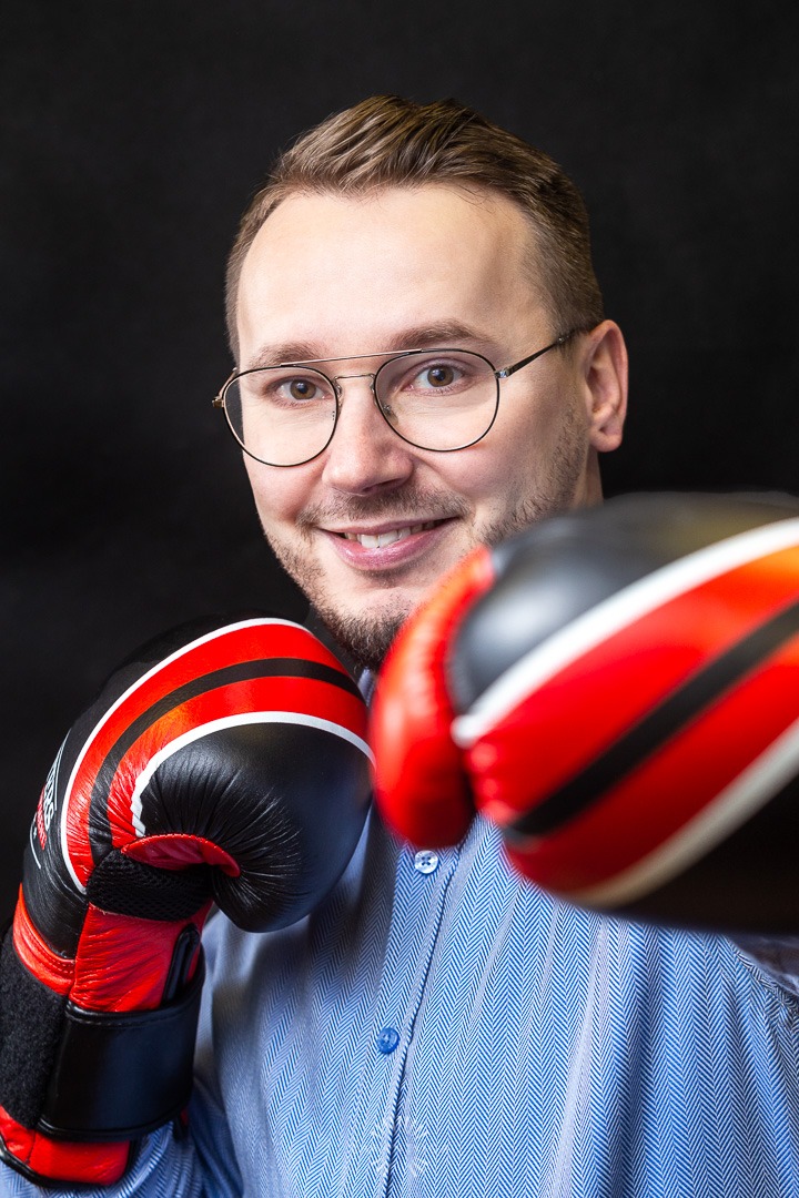 Portret uśmiechniętego mężczyzny w okularach, w niebieskiej koszuli w prążki, z czarno-czerwonymi rękawicami bokserskimi na tle ciemnego tła.