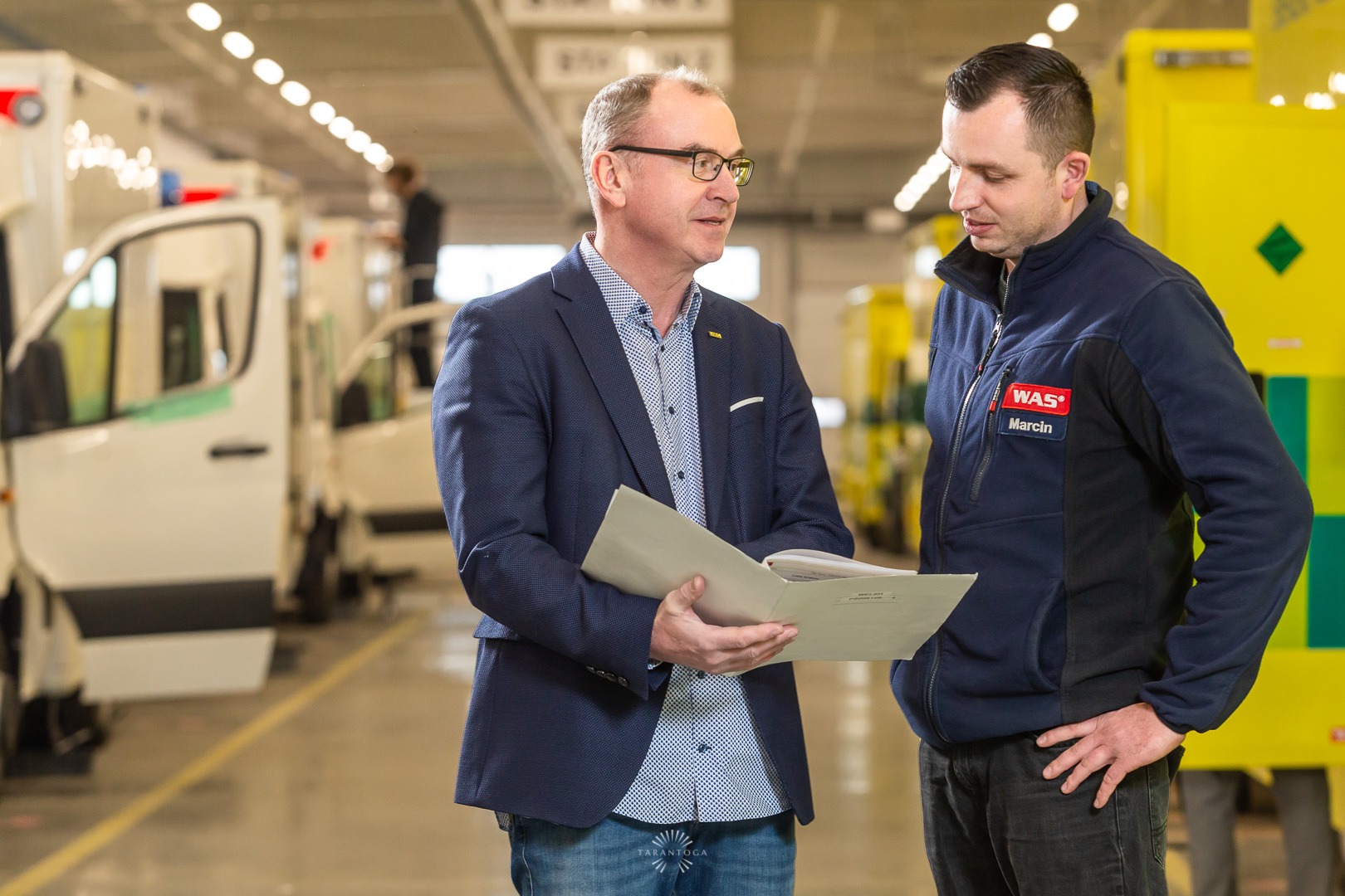 Dwaj mężczyźni w hali produkcyjnej ambulansów omawiają dokumenty, w tle widoczne pojazdy.