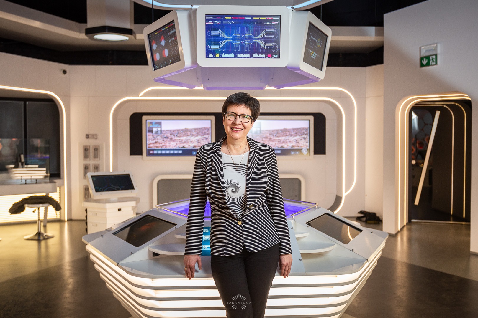 Portret kobiety w okularach stojącej w futurystycznym wnętrzu z monitorami i oświetleniem LED.