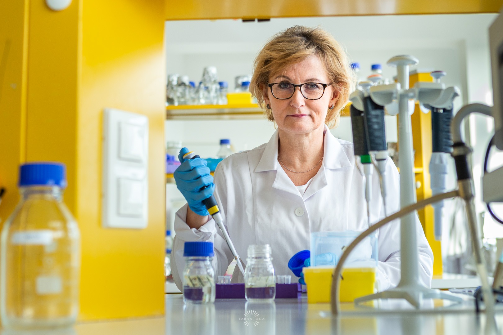 Kobieta w okularach i białym fartuchu laboratoryjnym, w niebieskich rękawicach, pipetuje ciecz do małej fiolki w laboratorium z żółtymi elementami wyposażenia.