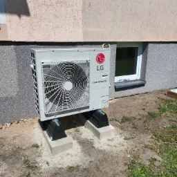 Zewnętrzna jednostka pompy ciepła LG Therma V zamontowana na betonowych podstawach przy ścianie budynku z oknem piwnicznym.