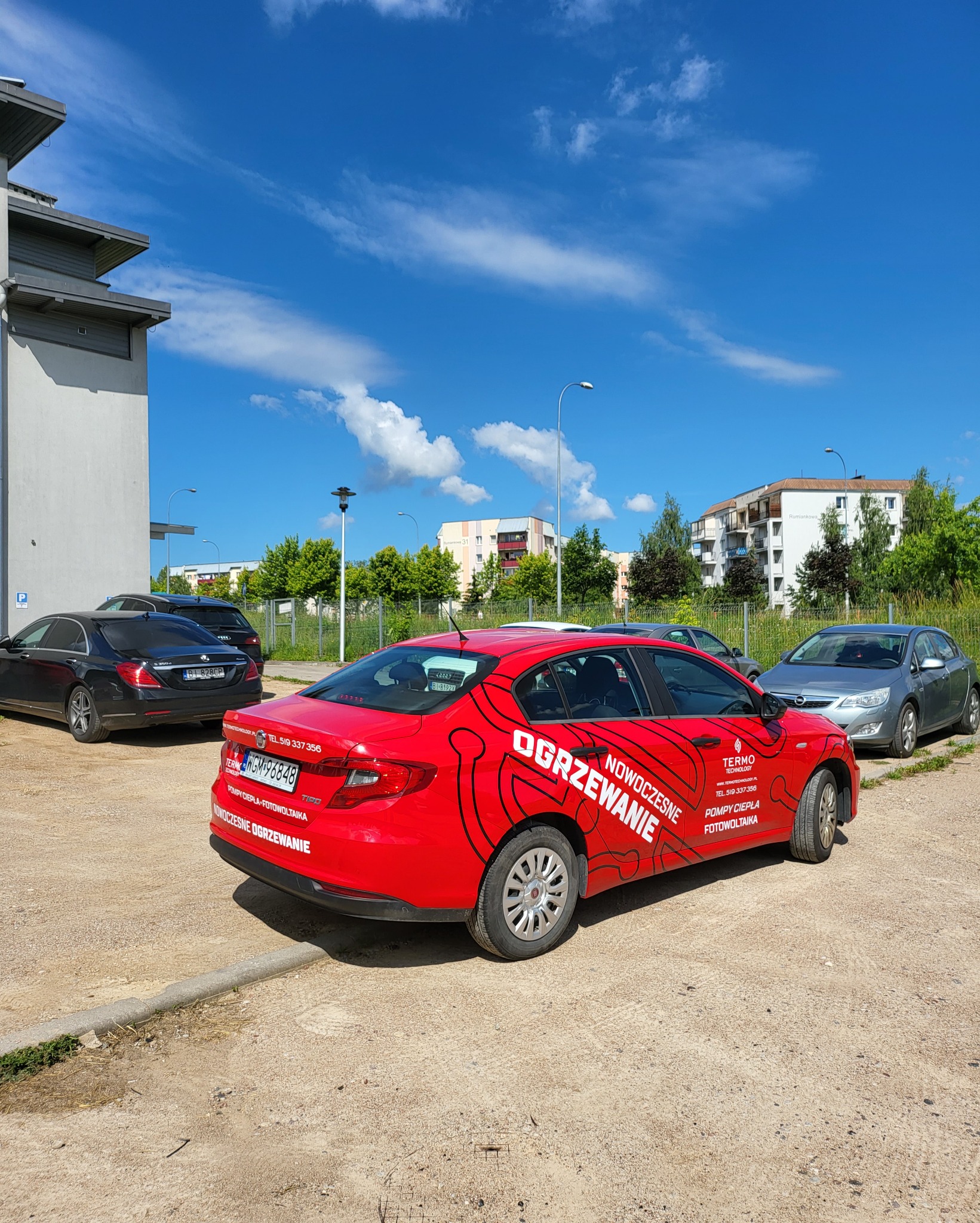 Czerwony samochód z reklamą usług grzewczych i fotowoltaicznych na parkingu w słoneczny dzień.