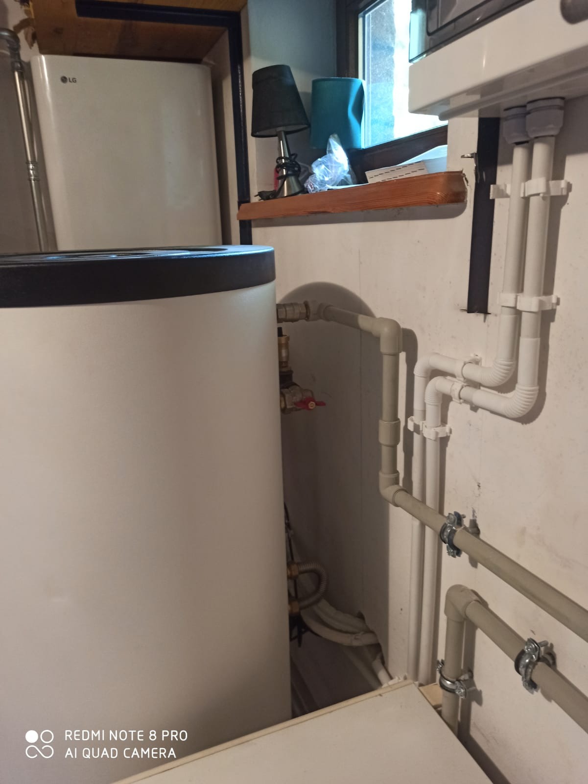 Biały zasobnik ciepłej wody użytkowej LG z widocznymi rurami instalacji grzewczej i zaworem bezpieczeństwa, umieszczony w pomieszczeniu z oknem i półką z lampką nocną.