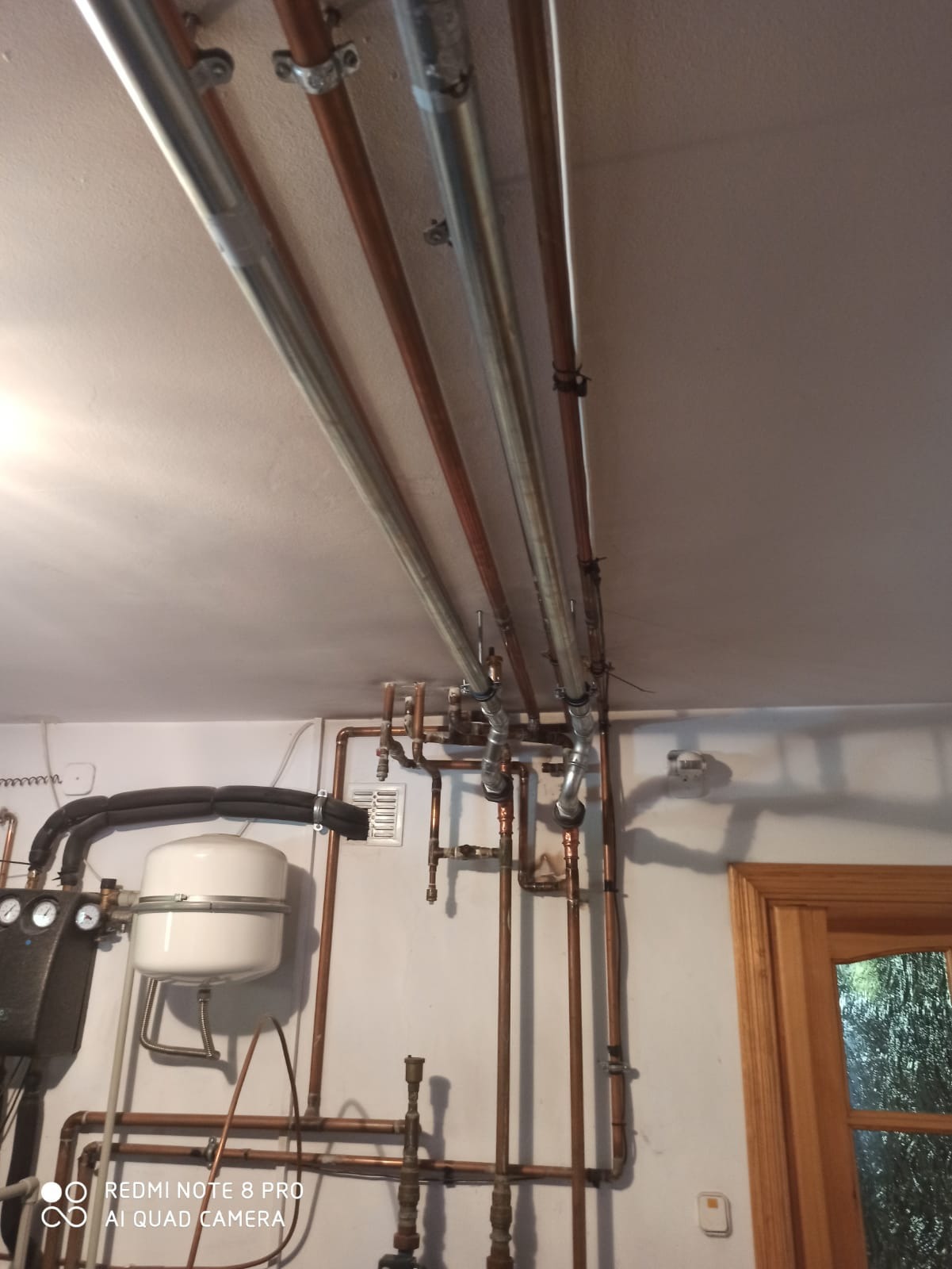 Widok instalacji grzewczej z rur miedzianych i stalowych, mocowanych do sufitu za pomocą obejm, z widocznym zbiornikiem wyrównawczym i fragmentem kotła oraz drewnianą ramą okienną.
