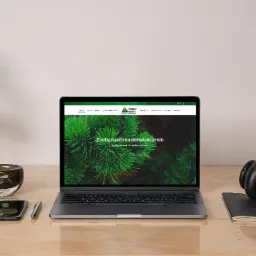 Laptop wyświetla stronę internetową szkółki drzewek i krzewów ozdobnych 'Green Market', obok leżą słuchawki, telefon i notatnik na blacie biurka.