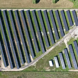 Widok z lotu ptaka na dużą farmę paneli słonecznych, z rzędami paneli rozciągającymi się na polu, mały budynek techniczny w rogu farmy.