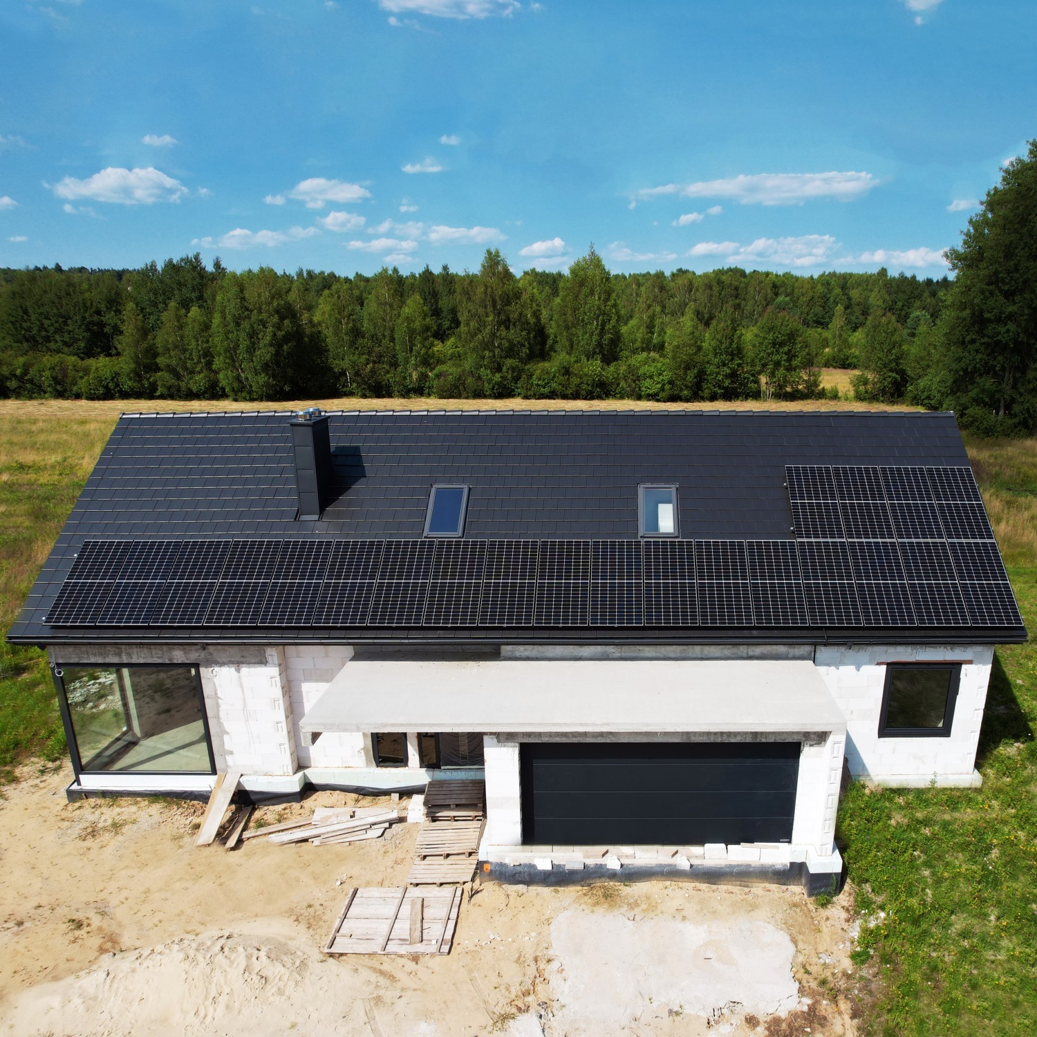 Niedokończony dom jednorodzinny z czarnym dachem pokrytym panelami słonecznymi, widok z góry na tle lasu i błękitnego nieba.