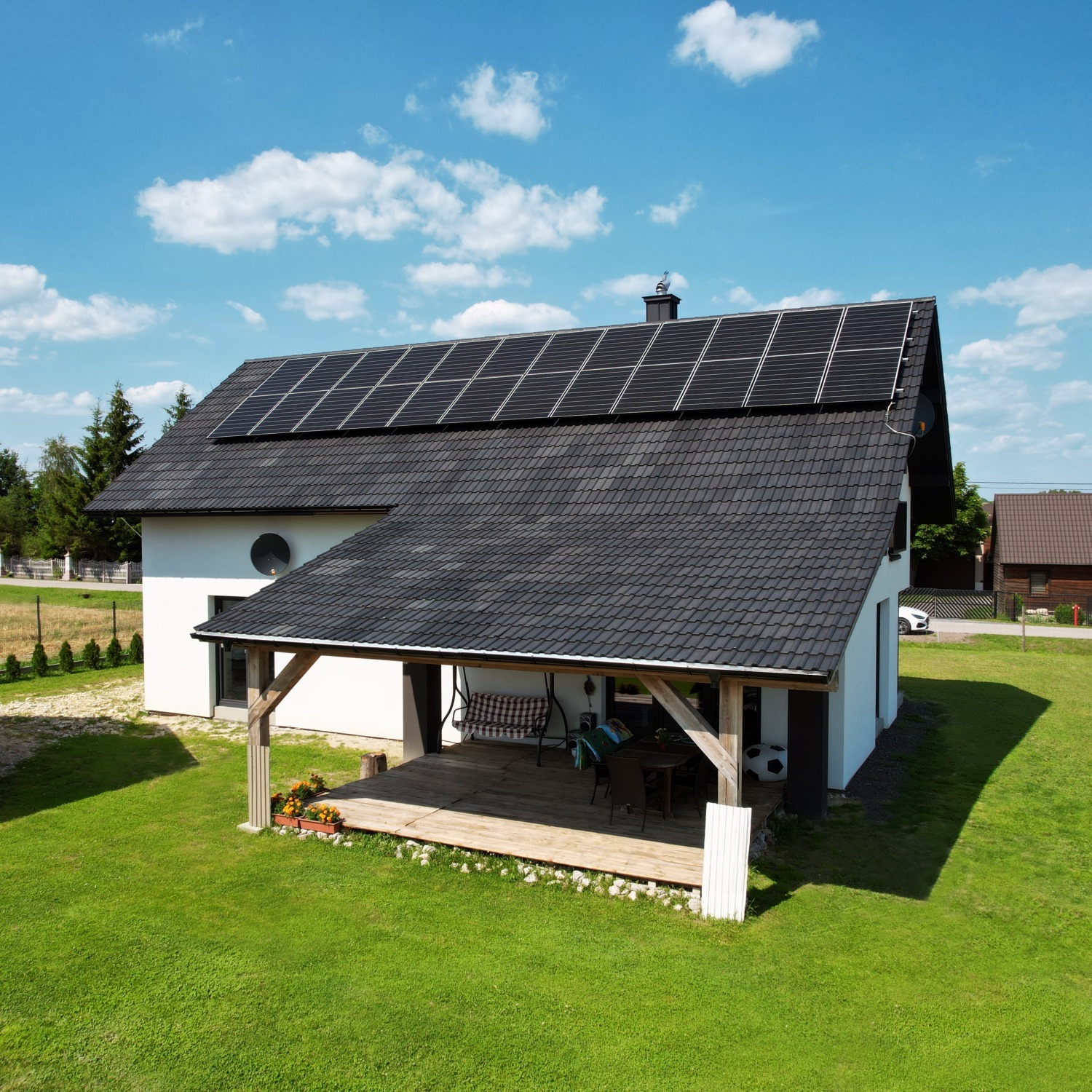 Biały dom jednorodzinny z ciemną dachówką i panelami słonecznymi na dachu, widok z góry, z drewnianym tarasem z huśtawką i meblami ogrodowymi, otoczony zielonym trawnikiem w słoneczny dzień.