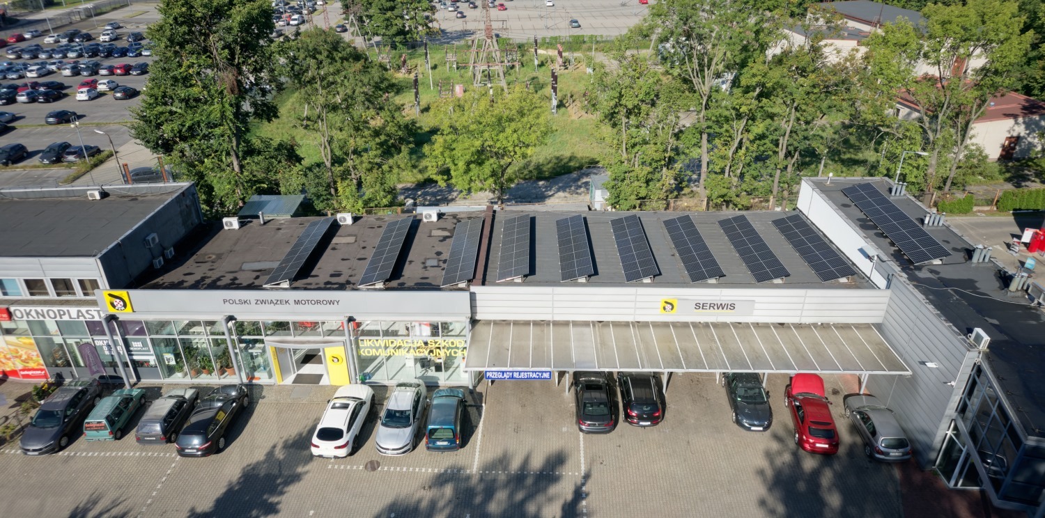 Widok z góry na budynek serwisu samochodowego z zainstalowanymi panelami fotowoltaicznymi na dachu, zaparkowanymi samochodami i otaczającą zielenią.