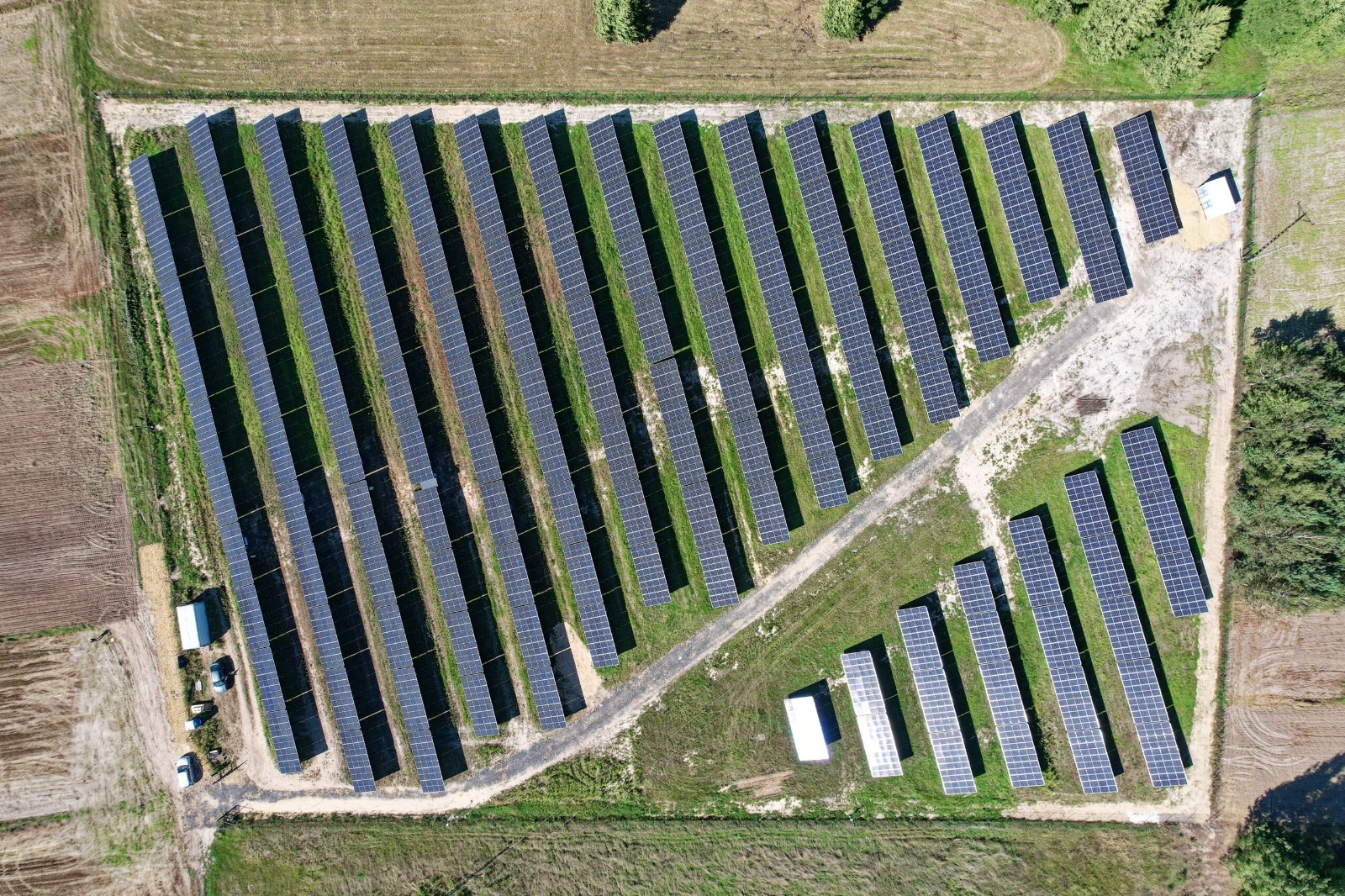 Widok z lotu ptaka na dużą farmę paneli słonecznych, z rzędami paneli rozciągającymi się na polu, mały budynek techniczny w rogu farmy.