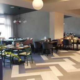 NOVA Technologie Obiektowe Sp.J. - Jasna, przestronna sala restauracyjna z parkietem ułożonym we wzór jodełki francuskiej, oświetlona kulistymi lampami, z rośliną doniczkową w lewym dolnym rogu.