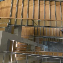 Widok z g&oacute;ry na drewniane schody z metalowymi wykończeniami i szklanymi balustradami, oświetlone światłem padającym z boku.