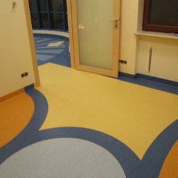 NOVA Technologie Obiektowe Sp.J. - Wielokolorowa podłoga z wykładziny PCV w kształcie fal, z widocznymi detalami łączeń i wzorów, w pomieszczeniu z oknem i drzwiami z matową szybą.