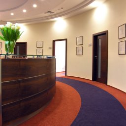 NOVA Technologie Obiektowe Sp.J. - Recepcja z zakrzywioną ladą z ciemnego drewna i wykładziną dywanową w kolorach czerwonym i granatowym, w tle białe ściany z ramkami i bukiet białych tulipanów.