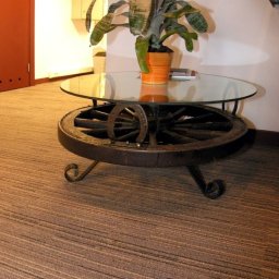 NOVA Technologie Obiektowe Sp.J. - Brązowa wykładzina dywanowa z subtelnym wzorem w paski, widoczny fragment szklanego stolika kawowego z podstawą z koła od wozu i rośliną doniczkową na blacie.