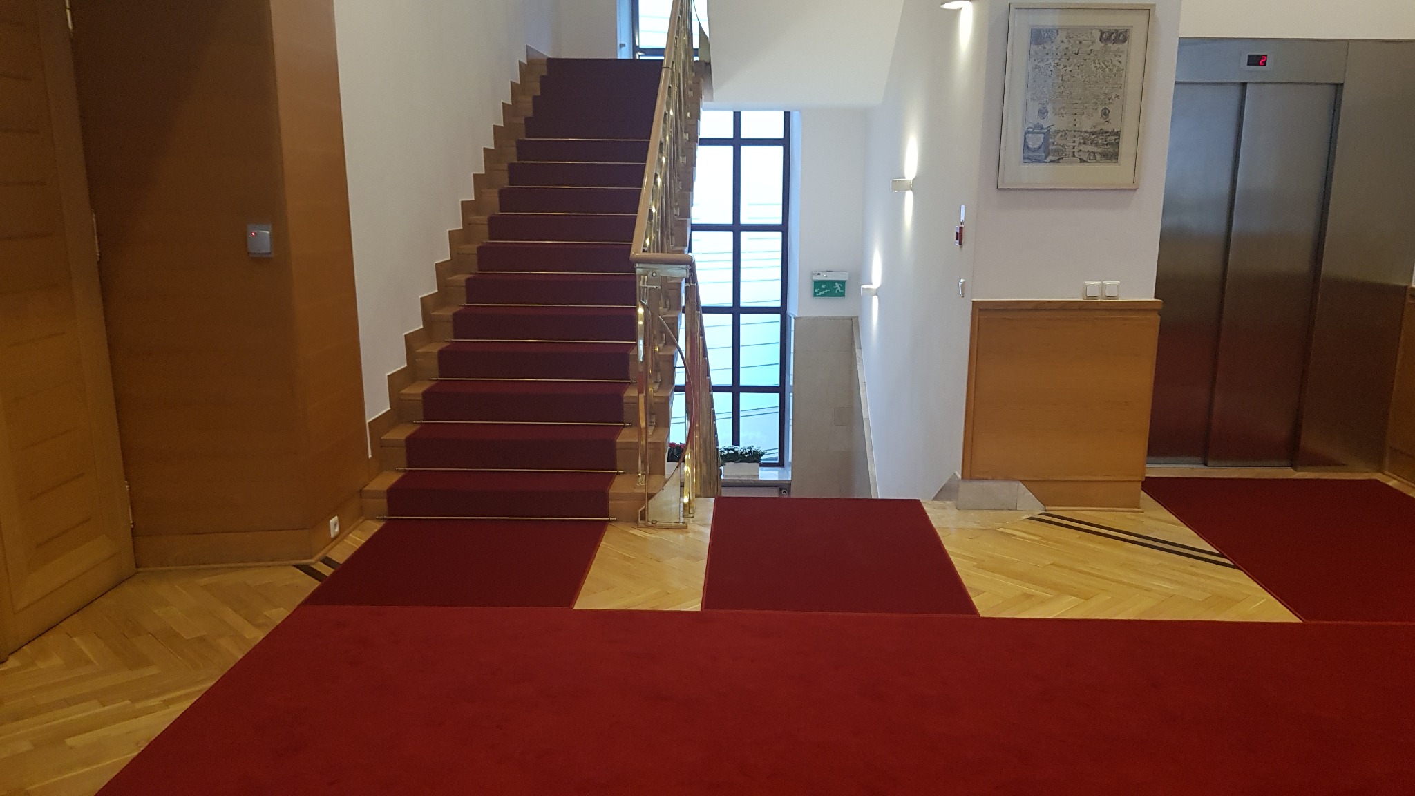Czerwona wykładzina dywanowa na schodach i podłodze w holu budynku z drewnianą podłogą parkietową, windą i oknem w tle.