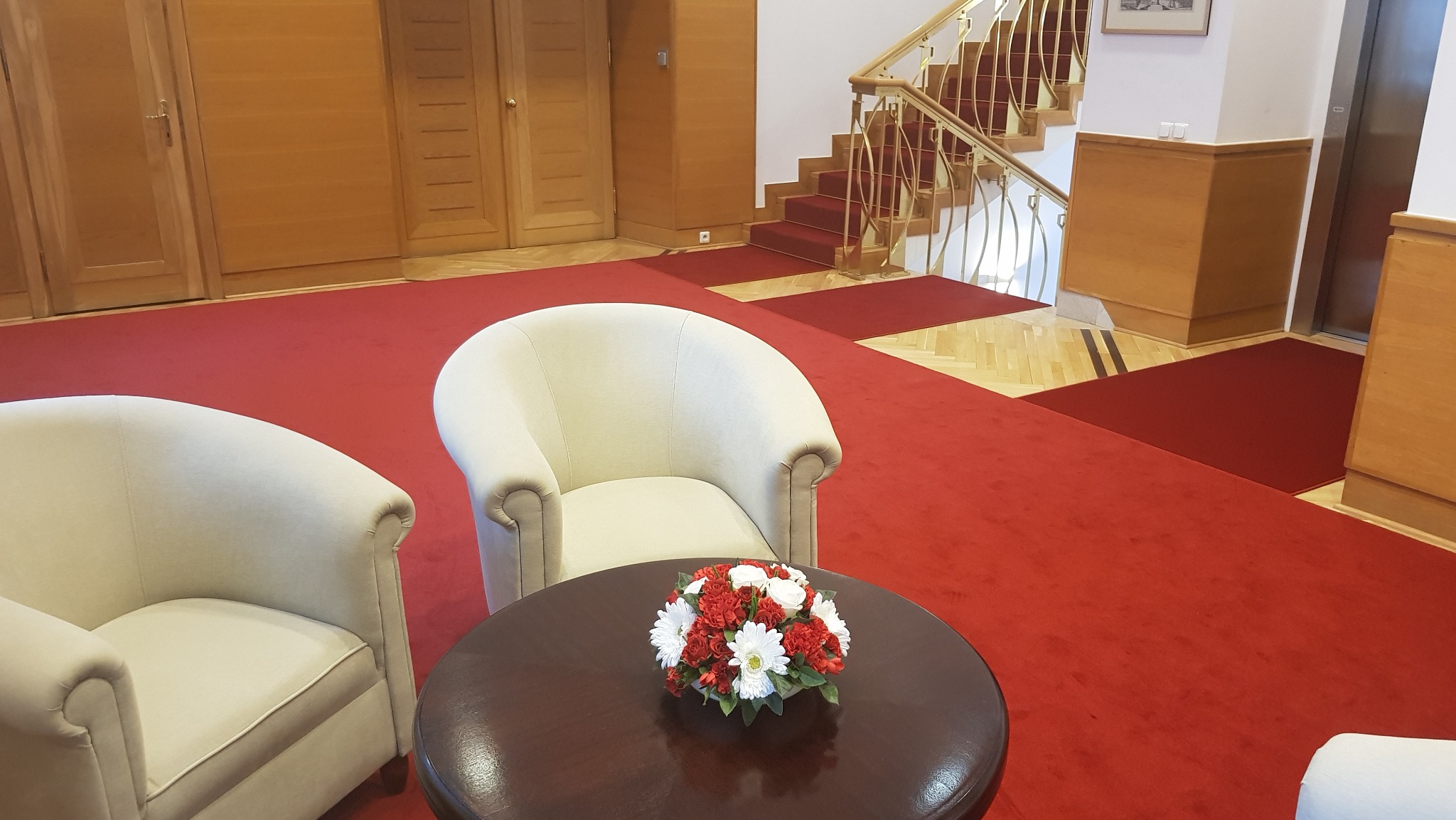 Czerwona wykładzina dywanowa w holu hotelowym z kremowymi fotelami i okrągłym stolikiem z kwiatami, schody z metalową balustradą w tle.