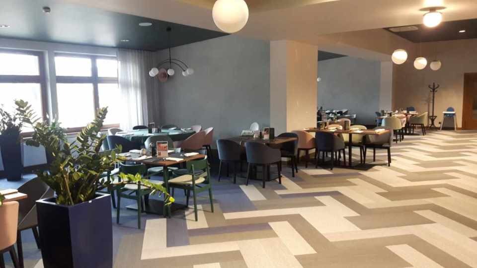 Jasna, przestronna sala restauracyjna z parkietem ułożonym we wzór jodełki francuskiej, oświetlona kulistymi lampami, z rośliną doniczkową w lewym dolnym rogu.