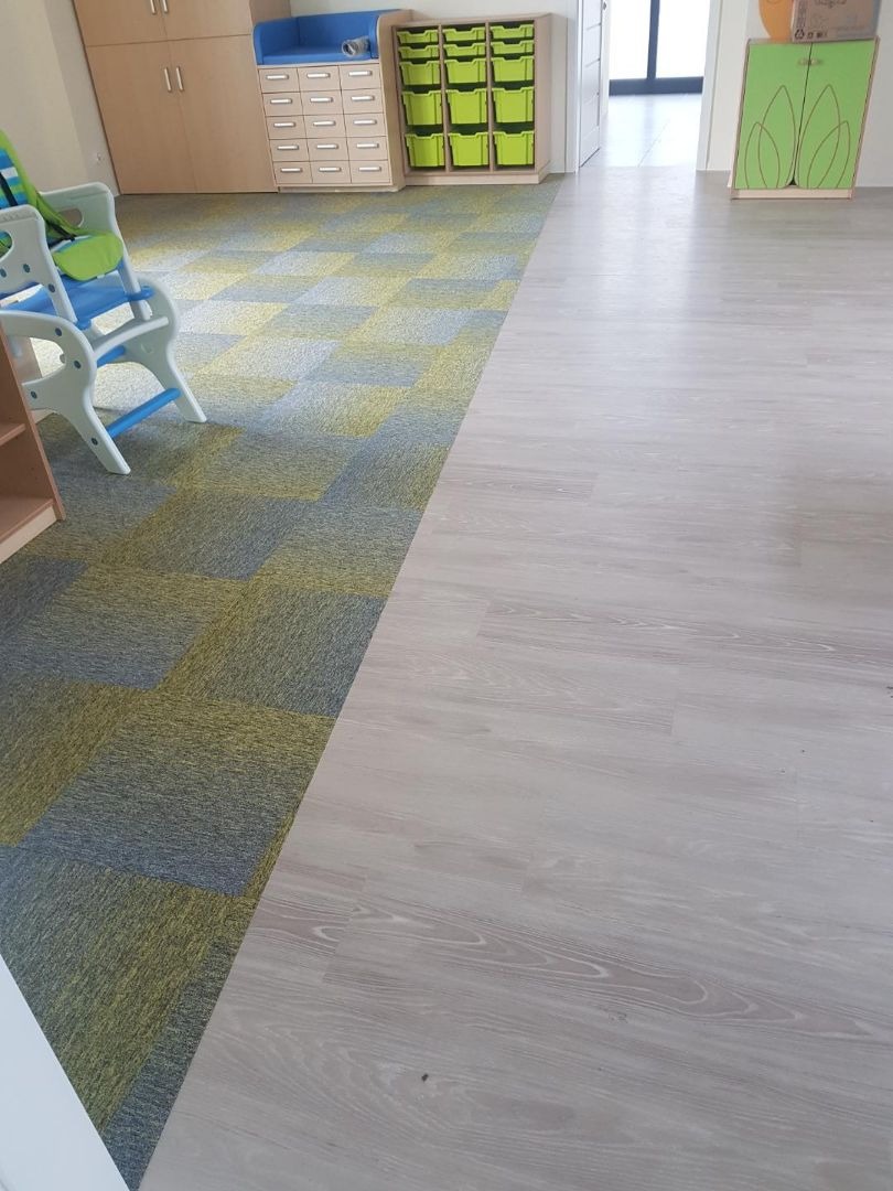Połączenie dwóch rodzajów podłogi w przedszkolu: szara wykładzina dywanowa w szachownicę przechodzi w panele podłogowe imitujące jasne drewno, widoczne meble dziecięce w tle.