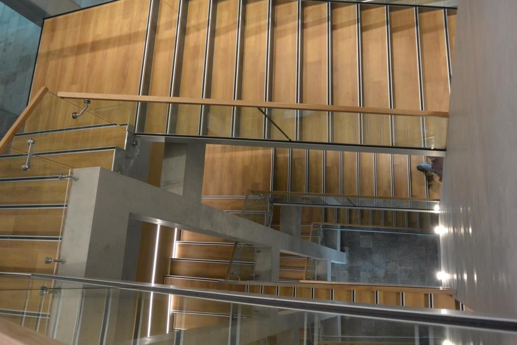 Widok z góry na drewniane schody z metalowymi wykończeniami i szklanymi balustradami, oświetlone światłem padającym z boku.