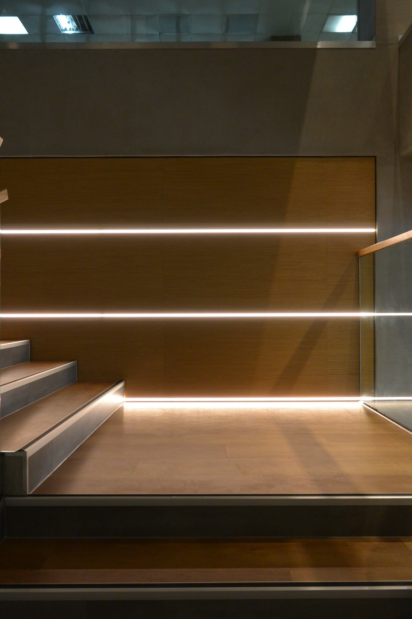 Schody z drewnianymi stopniami i podświetlaną drewnianą ścianą, szklana barierka po prawej stronie, minimalistyczny design.