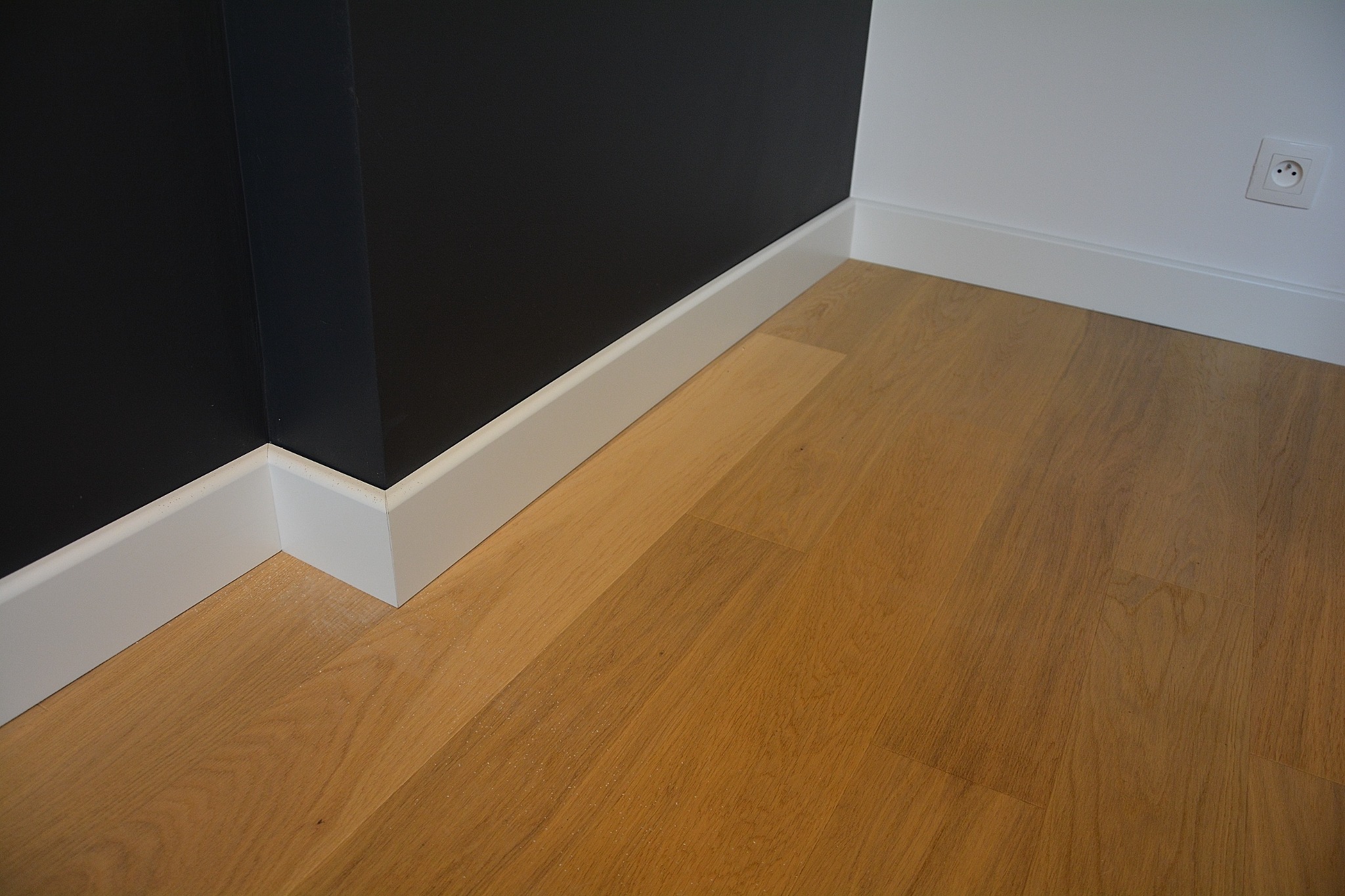 Jasna drewniana podłoga z białymi listwami przypodłogowymi na styku z czarną i białą ścianą, widoczny kontakt elektryczny.