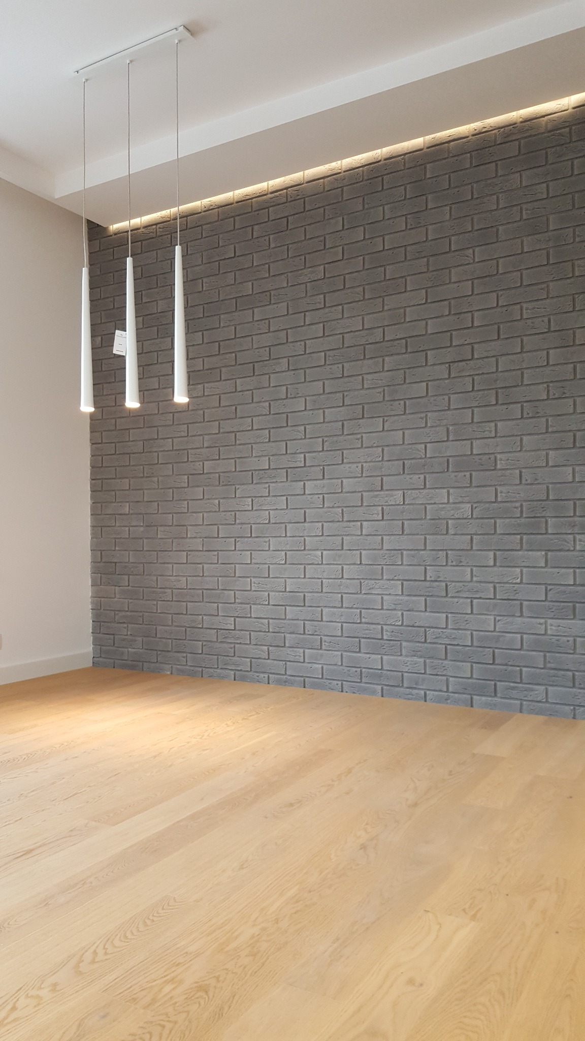 Jasna, lakierowana podłoga z drewna dębowego w pustym pokoju, z szarą ścianą z cegły i trzema wiszącymi lampami stożkowymi.