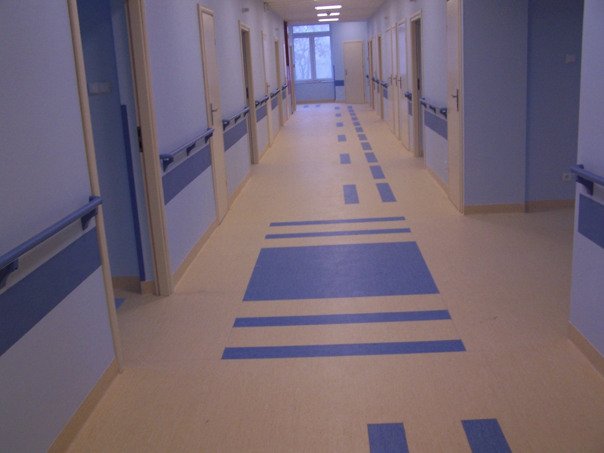 Długi korytarz w placówce medycznej z beżową podłogą z niebieskimi, geometrycznymi wzorami, jasnoniebieskie ściany z drewnianymi drzwiami i poręczami.